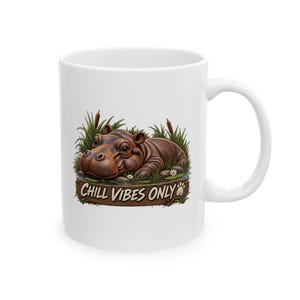 May include: A white ceramic mug featuring a graphic of a brown hippopotamus resting in a grassy pond with cattails and white flowers. Below the hippo, a wooden sign graphic displays the text CHILL VIBES ONLY with a small paw print icon.