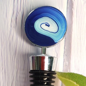 May include: A silver bottle stopper with a blue and white swirl design on the top. The stopper has a black rubber ring and a pointed tip.