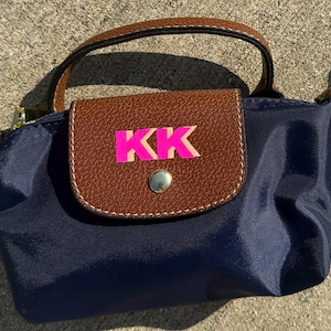 May include: Navy blue handbag with a brown leather flap and handle. The flap features the letters "KK" in pink. The bag has a small handle and a gold-colored clasp.