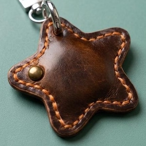 May include: A star-shaped dark brown leather keyring featuring thick orange stitching along the edges and a single brass-coloured metal rivet. The keyring is attached to a silver-toned metal lobster clasp and a split ring for keys.