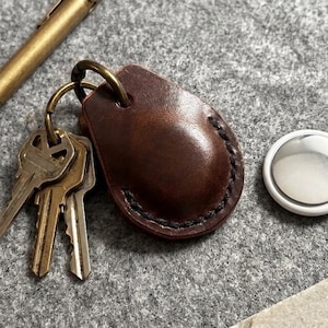 May include: A dark brown leather key fob with visible black stitching, attached to a brass key ring holding three metal keys. A brass pen lies nearby, and a white circular tracking device with a silver centre is positioned to the right.