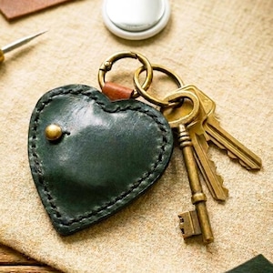 May include: A dark green heart-shaped leather keyring with visible stitching and a single brass stud. The keyring is attached to a brass ring holding two metal keys. A small white circular tracking device is positioned above the keyring.
