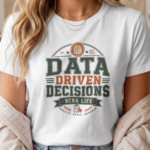 May include: A white short-sleeve t-shirt featuring a graphic design with the text "DATA DRIVEN DECISIONS" in dark green and orange. The design includes a brain icon, a clipboard with a puzzle piece, and the text "BCBA LIFE" and "ANALYZE. APPLY. EMPOWER."