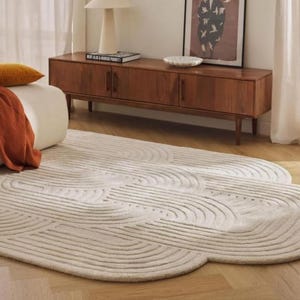 May include: A cream-colored, patterned rug with a cloud-like shape dominates the foreground. A mid-century modern wooden cabinet with three doors and a framed artwork are in the background. A white lamp and a decorative bowl sit on top of the cabinet.