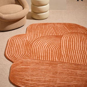 May include: A distinctive orange and cream-coloured rug with an irregular shape. The rug displays a pattern of curved lines, giving it a textured look. It's positioned on a neutral floor, with a beige chair and side table visible in the background.