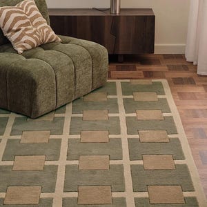 May include: A modern area rug with a geometric pattern in shades of green and beige. The rug features a repeating rectangular design, complementing the olive green upholstered furniture and zebra print pillow in the room. The rug is a stylish addition to any living space.