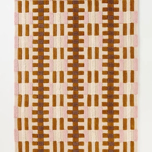 Puede incluir: Una alfombra rectangular con un patrón geométrico repetitivo en tonos marrón, beige y rosa. La alfombra tiene un borde ondulado en un color marrón más oscuro. El diseño presenta bloques verticales y horizontales, creando una apariencia texturizada.