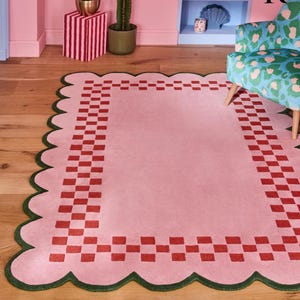 Op de afbeelding: Een roze rechthoekig vloerkleed met een geschulpte rand en een rode dambordrand. Het vloerkleed heeft een groene rand en ligt op een houten vloer. Het vloerkleed ligt in een kamer met roze muren en een stoel met patroon.
