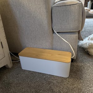 May include: A rectangular cable management box featuring a solid white plastic base and a removable light brown bamboo wood lid. The container is designed to conceal power strips and electrical cords, with a white cord exiting from the side.