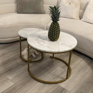 May include: Two round coffee tables with white marble tops and gold metal frames. A pineapple sits on one table. A white cat is under one table. The tables are in front of a cream-colored sofa.