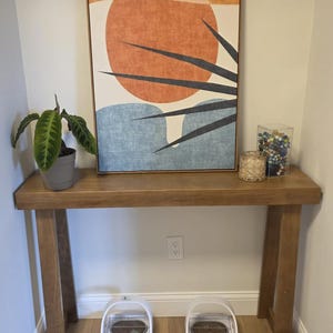 May include: A rectangular framed wall art piece featuring an abstract sun and palm leaf design in orange, blue, and cream. The artwork is displayed on a wooden console table alongside a potted plant, a candle, and a clear container of marbles.