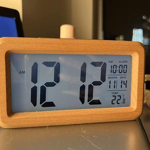 May include: Digital alarm clock with a light wood frame displaying the time as 12:12 AM. The screen also shows the alarm time, date, and temperature in Celsius. The clock has a simple, modern design.