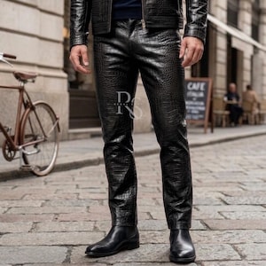 Può includere: Pantaloni in pelle nera con texture a pelle di coccodrillo. I pantaloni sono indossati con stivali in pelle nera e una giacca abbinata. I pantaloni hanno un taglio dritto e una chiusura con cerniera. L'immagine è scattata all'aperto su una strada acciottolata.