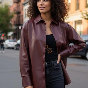 以下が含まれることがあります： 長袖のバーガンディ色のボタンダウンシャツ。 シャツは革のような素材でできており、黒のジーンズと黒のタンクトップと組み合わせています。 画像は、都市の背景を背景に屋外で撮影されています。