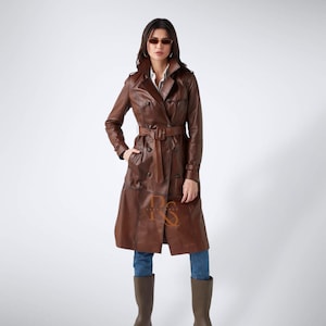 Könnte beinhalten: Ein brauner Ledertrenchcoat mit doppelreihigem Design, Gürtel in der Taille und klassischem Kragen. Der Mantel wird mit blauen Jeans und braunen Stiefeln getragen.