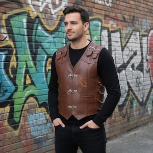 May include: Brown leather vest with silver-colored clasps and buckles. The vest has a V-neck and is worn over a black long-sleeve shirt. The background is a brick wall covered in colorful graffiti.