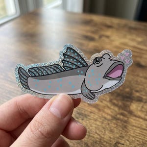 May include: A sticker featuring a cartoon fish with a gray body, blue spots, and a wide-open mouth. The fish has gray and blue fins and a silver glitter border. Pink hearts are near the fish's head.