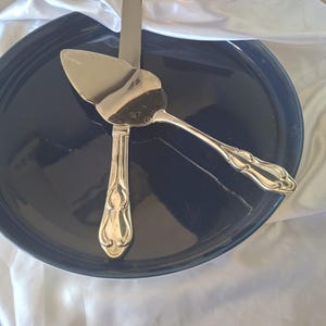 May include: A silver cake server and knife set on a dark blue plate. The server has a triangular blade and decorative handle. The knife is straight-edged with a matching handle. The set rests on a white cloth.