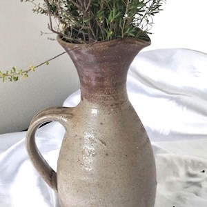 May include: A tall, handcrafted ceramic pitcher with a speckled beige body and a darker, brownish-red glazed neck. The vessel features a curved handle on the side and a flared rim, currently holding a bundle of thin, leafy green branches.