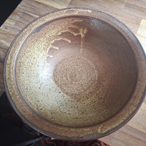 May include: A round ceramic bowl with a wide rim, featuring a speckled brown and tan glaze. The interior surface displays a textured, mottled appearance with darker brown tones concentrated at the base and lighter, sandy hues towards the edges.