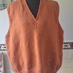 以下が含まれることがあります： Vネックのデザインの、オレンジブラウンのニットベスト。ノースリーブの衣服は黒いマネキンに展示されています。ベストはリブ編みの裾とネックラインを持っています。ニットウェアは無地で、柔らかい糸で作られているようです。