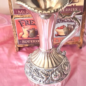 May include: A silver-toned metal pitcher featuring a flared, scalloped rim and a curved handle. The body displays a smooth, fluted neck above a wide, rounded base decorated with a detailed embossed floral pattern of roses and leaves.