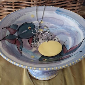 May include: A decorative ceramic pedestal bowl with a blue and white watercolour design. The bowl features a painted fish motif in teal, red, and black. Silver necklaces and a yellow pendant rest inside the bowl. The rim is dotted with yellow accents.