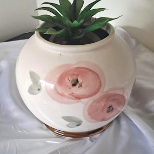 May include: A round, cream-coloured ceramic planter featuring hand-painted pink floral motifs and small grey leaves. The base of the planter has a brown glazed rim. A green artificial succulent plant is placed inside the opening of the container.