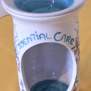 May include: A white ceramic oil burner with a cylindrical body and a wide, flared top rim. The interior of the top bowl and the base are painted a solid blue. The text "ESSENTIAL CARE" is written in blue script above a large, arched opening.