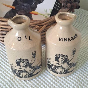May include: Two beige ceramic cruets for oil and vinegar, each featuring a black graphic illustration of a cherub with a wine barrel and a goblet. The bottles have a cylindrical body, a narrow neck, and a small pouring spout at the top rim.