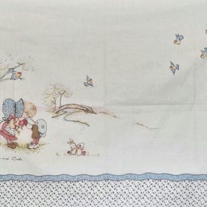 May include: A white fabric panel featuring a printed illustration of two children, labelled "Lucy and Luke", standing under a tree. The scene includes small birds, a bridge, and a floral border with a blue scalloped trim and a small floral pattern.