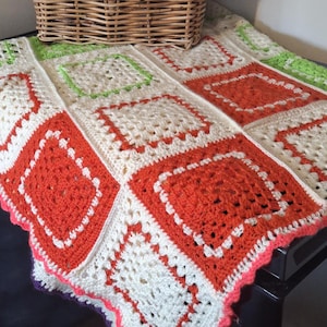 May include: A handmade crochet blanket with a patchwork design. The blanket features cream, orange, and green squares, with a pink scalloped border. A woven basket is in the background, adding a rustic touch to the cozy, colourful textile.