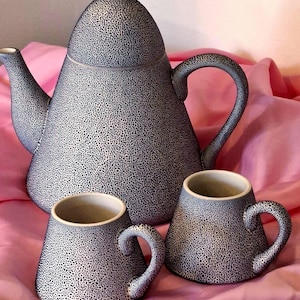 May include: A ceramic tea set consisting of a large teapot with a lid and two matching mugs. Each piece features a textured, speckled pattern in black and white, with a smooth, light-coloured interior finish on the mugs and a curved handle.