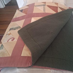 May include: A quilted blanket featuring a geometric patchwork top in shades of beige, tan, and muted rose with a textured surface. The underside of the blanket is a solid, dark olive green felted material with a stitched border along the edges.