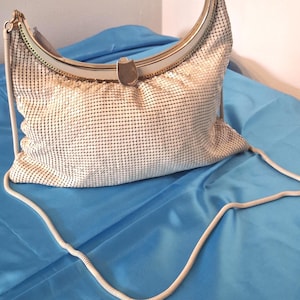 May include: A cream-coloured metal mesh handbag featuring a curved top frame with a metallic clasp closure. The bag includes a long, thin, matching cream-coloured shoulder strap attached to the sides of the frame, creating a crescent-shaped silhouette.