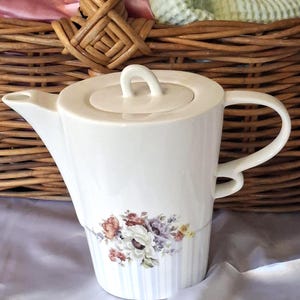 May include: A white ceramic teapot featuring a cylindrical body with a subtle vertical striped pattern and a floral bouquet decal in shades of red, purple, and yellow. The teapot includes a matching lid with a small handle, a spout, and a curved handle.