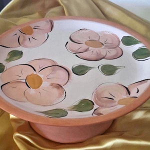 May include: A round ceramic pedestal plate with a terracotta-coloured rim and base. The flat surface features a cream-coloured background decorated with hand-painted light pink flowers with yellow centres and small green leaves scattered throughout.