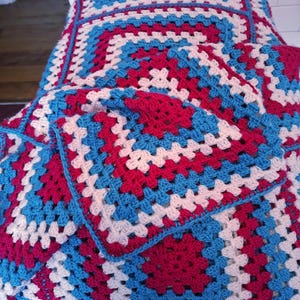May include: A handmade crochet blanket featuring a repeating granny square pattern. The textile is composed of alternating rows of yarn in vibrant shades of magenta, bright turquoise, and off-white, creating a geometric, multi-coloured design.