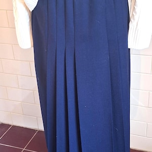 May include: A solid navy blue skirt featuring a high-waisted design with vertical knife pleats that extend from the waistband to the hem. The skirt is paired with a white long-sleeved jumper that has a visible cable knit texture on the sleeves.