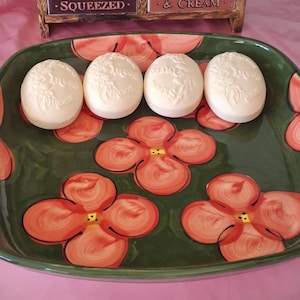 May include: A green, rectangular ceramic serving dish with a floral design, holding four oval, off-white soaps. The dish has a dark green base with large orange flowers and a pink border. The soaps have embossed floral details.