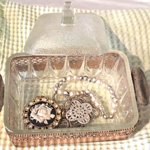 May include: A square clear glass trinket box with a textured lid and a metallic base. Inside the box sits a gold-toned brooch featuring a white rose cameo surrounded by rhinestones, alongside a bracelet made of white pearls and silver beads.