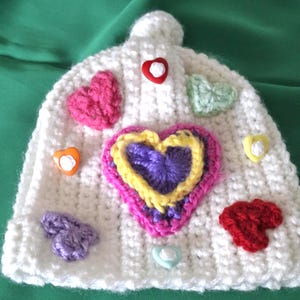 May include: A white crocheted beanie featuring a large central heart motif layered in purple, yellow, and pink yarn. The hat is decorated with several smaller crocheted hearts in pink, red, purple, and light green, along with small heart-shaped buttons.