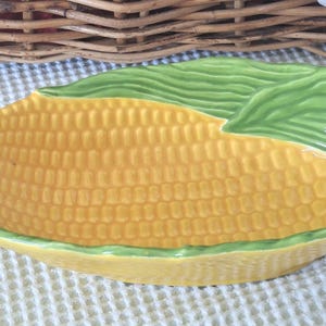 May include: A ceramic serving dish shaped like an ear of corn. The dish is yellow with a textured surface to mimic corn kernels, and green leaves at the top. The dish is on a white textured surface.