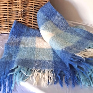 May include: A soft, felted wool throw blanket featuring a blue and white checkered pattern. The blanket has a textured, fuzzy surface and is finished with long, loose fringe along the edges, displaying various shades of light and dark blue.