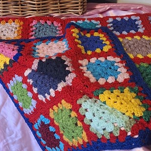 May include: A colourful, handmade crochet blanket with a patchwork design. The blanket has a red background with squares in blue, yellow, green, pink, and white. The blanket is made of yarn and has a textured appearance.
