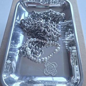 May include: A silver-toned rectangular tray containing a collection of jewellery, including a thick silver chain and a bracelet featuring white faux pearls, small silver beads, and a four-leaf clover charm encrusted with clear sparkling stones.