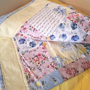 May include: A quilted fabric cover featuring a patchwork design of floral patterns in shades of blue, pink, and yellow. The piece includes a solid pale yellow border and is secured with three round buttons in white and light brown colors.