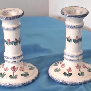 May include: A pair of off-white ceramic candle holders featuring a hand-painted floral pattern with pink tulip-like flowers and green leaves. The rims and bases are decorated with a textured blue sponge-painted trim, creating a consistent design.