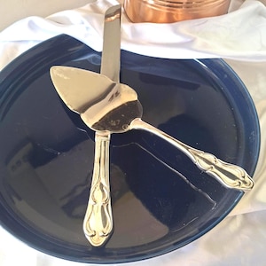 May include: A silver-toned cake server and a matching knife with ornate, embossed handles. The utensils are arranged in a cross pattern, resting on a smooth, dark blue circular plate. The metal surfaces are polished and reflective.