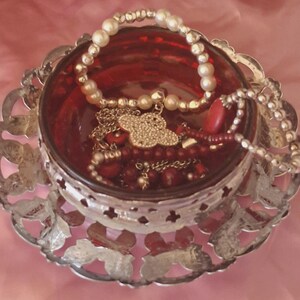 May include: A deep red glass bowl set within a decorative silver-toned metal filigree holder. The bowl contains a collection of jewellery, including a white pearl-style beaded bracelet, a gold-toned clover charm necklace, and red beaded strands.